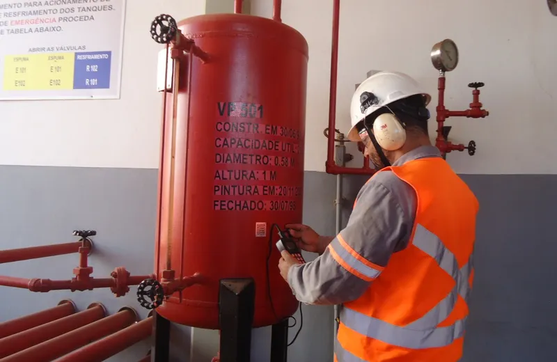 Técnico com colete de segurança laranja inspecionando vaso de pressão vermelho VP 501